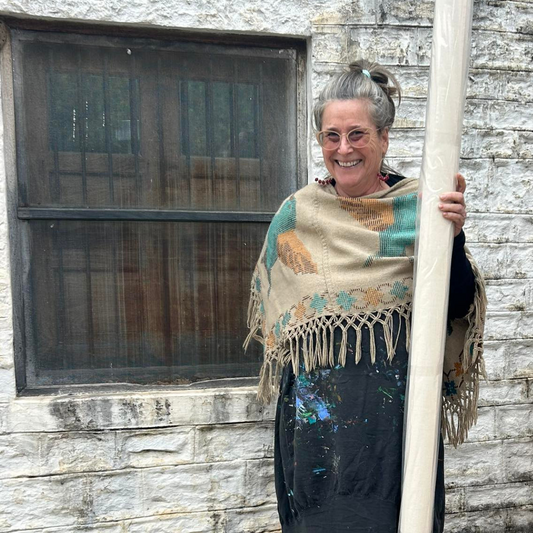 Lisa Regan, Tulsa-based metal sculptor and abstract painter, standing outside her studio, holding a rolled canvas, dressed in a paint-splattered apron and a cozy shawl, radiating her creative spirit.