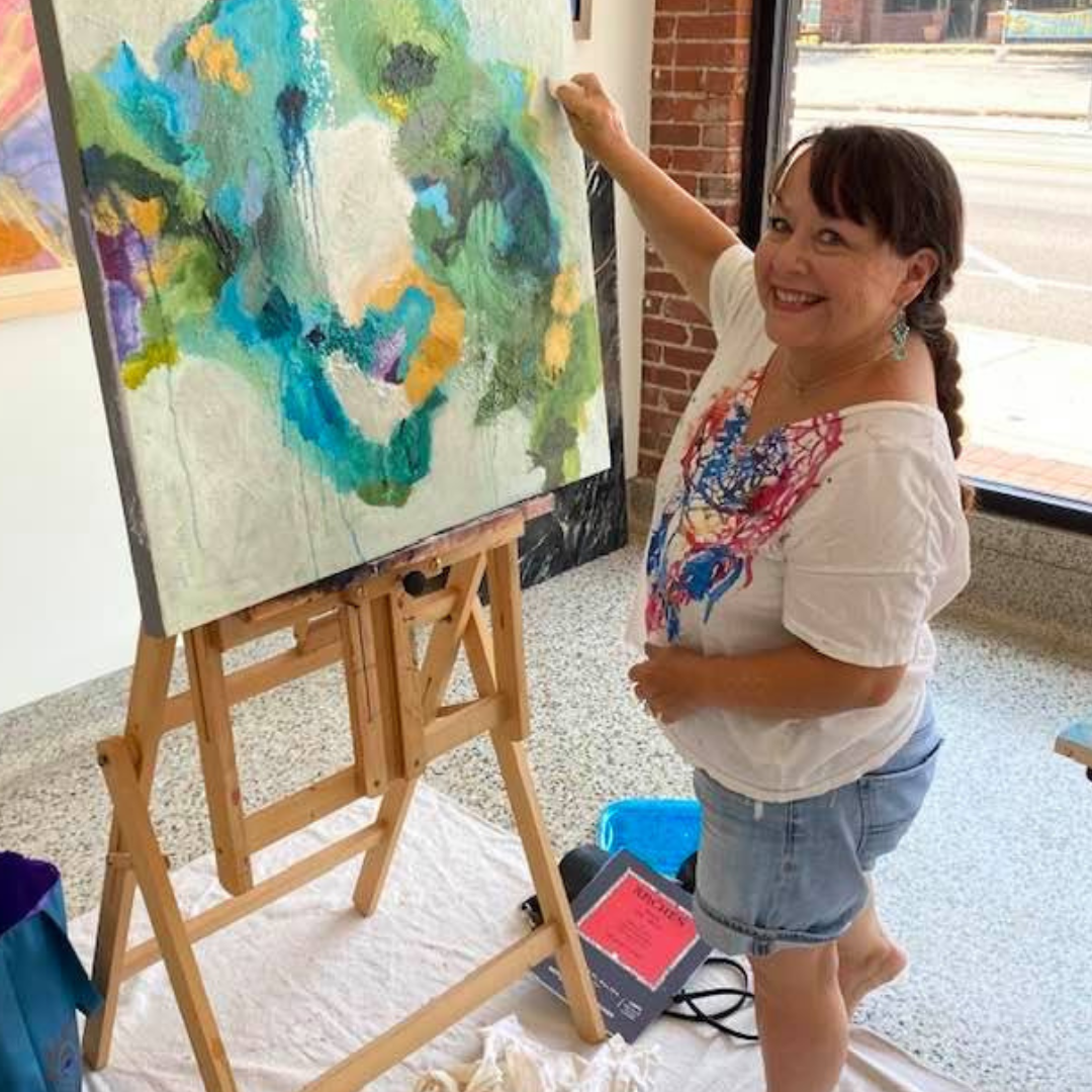 Pam Watson, a Tulsa-based abstract artist, smiles while painting a vibrant, textured piece on an easel, showcasing her creative process in a sunny art studio.